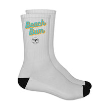 Ein Paar umweltfreundliche Unisex-Crew-Socken in Weiß mit schwarzer Spitze und Knöchelteil und einem kleinen Logo und Text im Retro-Stil "Beach Bum". Die Socken haben ein sportliches Design und eine bequeme Passform mit gepolstertem Fußbett. Die verwendeten Materialien sind umweltverträglich und fördern einen bewussten Konsum. Die Socken sind sowohl für Männer als auch für Frauen geeignet und können bei verschiedenen Aktivitäten getragen werden.