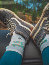 Ein Paar umweltfreundliche Unisex-Crew-Socken in Weiß mit schwarzer Spitze und Knöchelteil und einem kleinen Logo und Text im Retro-Stil "Beach Bum". Die Socken haben ein sportliches Design und eine bequeme Passform mit gepolstertem Fußbett. Die verwendeten Materialien sind umweltverträglich und fördern einen bewussten Konsum. Die Socken sind sowohl für Männer als auch für Frauen geeignet und können bei verschiedenen Aktivitäten getragen werden.