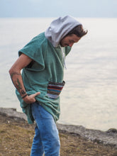 Eine Person, die einen bunten Boho Hippie Surf-Poncho mit aztekischem Muster trägt, steht an einem Strand. Der Poncho reicht bis knapp über die Knie und scheint aus einem dicken, saugfähigen Material zu bestehen. Bohemian Tribal Ethnic Patterned Colorful Beachwear Surfing Hooded Poncho Oversized Unisex Hippie-chic Festival fashion Beach accessory Mexican-inspired Southwestern Geometric Vibrant Multicolored Soft cotton Beach towel Quick-drying surfumkleide