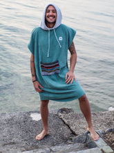 Eine Person, die einen bunten Boho Hippie Surf-Poncho mit aztekischem Muster trägt, steht an einem Strand. Der Poncho reicht bis knapp über die Knie und scheint aus einem dicken, saugfähigen Material zu bestehen. Bohemian Tribal Ethnic Patterned Colorful Beachwear Surfing Hooded Poncho Oversized Unisex Hippie-chic Festival fashion Beach accessory Mexican-inspired Southwestern Geometric Vibrant Multicolored Soft cotton Beach towel Quick-drying surfumkleide