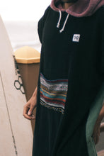 Eine Person, die einen bunten Boho Hippie Surf-Poncho mit aztekischem Muster trägt, steht an einem Strand. Der Poncho reicht bis knapp über die Knie und scheint aus einem dicken, saugfähigen Material zu bestehen. Bohemian Tribal Ethnic Patterned Colorful Beachwear Surfing Hooded Poncho Oversized Unisex Hippie-chic Festival fashion Beach accessory Mexican-inspired Southwestern Geometric Vibrant Multicolored Soft cotton Beach towel Quick-drying surfumkleide