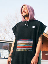 Eine Person, die einen bunten Boho Hippie Surf-Poncho mit aztekischem Muster trägt, steht an einem Strand. Der Poncho reicht bis knapp über die Knie und scheint aus einem dicken, saugfähigen Material zu bestehen. Bohemian Tribal Ethnic Patterned Colorful Beachwear Surfing Hooded Poncho Oversized Unisex Hippie-chic Festival fashion Beach accessory Mexican-inspired Southwestern Geometric Vibrant Multicolored Soft cotton Beach towel Quick-drying surfumkleide