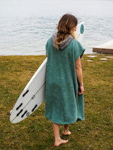 A person wearing a colorful boho hippie surf poncho with an aztec pattern stands on a beach. The poncho comes down to just above the knees and appears to be made out of a thick, absorbent material. Bohemian Tribal Ethnic Patterned Colorful Beachwear Surfing Hooded Poncho Oversized Unisex Hippie-chic Festival fashion Beach accessory Mexican-inspired Southwestern Geometric Vibrant Multicolored Soft cotton Beach towel Quick-drying surf changing room