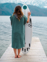 Eine Person, die einen bunten Boho Hippie Surf-Poncho mit aztekischem Muster trägt, steht an einem Strand. Der Poncho reicht bis knapp über die Knie und scheint aus einem dicken, saugfähigen Material zu bestehen. Bohemian Tribal Ethnic Patterned Colorful Beachwear Surfing Hooded Poncho Oversized Unisex Hippie-chic Festival fashion Beach accessory Mexican-inspired Southwestern Geometric Vibrant Multicolored Soft cotton Beach towel Quick-drying surfumkleide