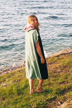 Eine Person, die einen bunten Boho Hippie Surf-Poncho mit aztekischem Muster trägt, steht an einem Strand. Der Poncho reicht bis knapp über die Knie und scheint aus einem dicken, saugfähigen Material zu bestehen. Bohemian Tribal Ethnic Patterned Colorful Beachwear Surfing Hooded Poncho Oversized Unisex Hippie-chic Festival fashion Beach accessory Mexican-inspired Southwestern Geometric Vibrant Multicolored Soft cotton Beach towel Quick-drying surfumkleide