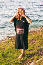 Eine Person, die einen bunten Boho Hippie Surf-Poncho mit aztekischem Muster trägt, steht an einem Strand. Der Poncho reicht bis knapp über die Knie und scheint aus einem dicken, saugfähigen Material zu bestehen. Bohemian Tribal Ethnic Patterned Colorful Beachwear Surfing Hooded Poncho Oversized Unisex Hippie-chic Festival fashion Beach accessory Mexican-inspired Southwestern Geometric Vibrant Multicolored Soft cotton Beach towel Quick-drying surfumkleide