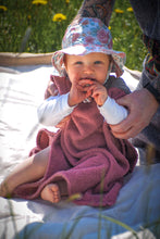 A baby sits on the beach wearing a colorful surf poncho with an Aztec-inspired boho pattern. The poncho is made of a soft, absorbent material that keeps baby warm and dry after swimming. The vibrant and multicolored pattern features ethnic details that give it a hippie-chic feel. The surf poncho has a hood to protect baby's head from the wind so that it is fully covered up to baby's knees. Boho hippie aztec