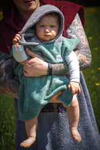 A baby sits on the beach wearing a colorful surf poncho with an Aztec-inspired boho pattern. The poncho is made of a soft, absorbent material that keeps baby warm and dry after swimming. The vibrant and multicolored pattern features ethnic details that give it a hippie-chic feel. The surf poncho has a hood to protect baby's head from the wind so that it is fully covered up to baby's knees. Boho hippie aztec