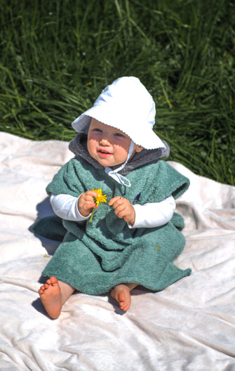 A baby sits on the beach wearing a colorful surf poncho with an Aztec-inspired boho pattern. The poncho is made of a soft, absorbent material that keeps baby warm and dry after swimming. The vibrant and multicolored pattern features ethnic details that give it a hippie-chic feel. The surf poncho has a hood to protect baby's head from the wind so that it is fully covered up to baby's knees. Boho hippie aztec