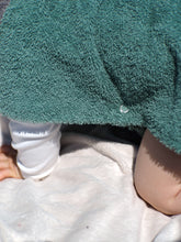 A baby sits on the beach wearing a colorful surf poncho with an Aztec-inspired boho pattern. The poncho is made of a soft, absorbent material that keeps baby warm and dry after swimming. The vibrant and multicolored pattern features ethnic details that give it a hippie-chic feel. The surf poncho has a hood to protect baby's head from the wind so that it is fully covered up to baby's knees. Boho hippie aztec